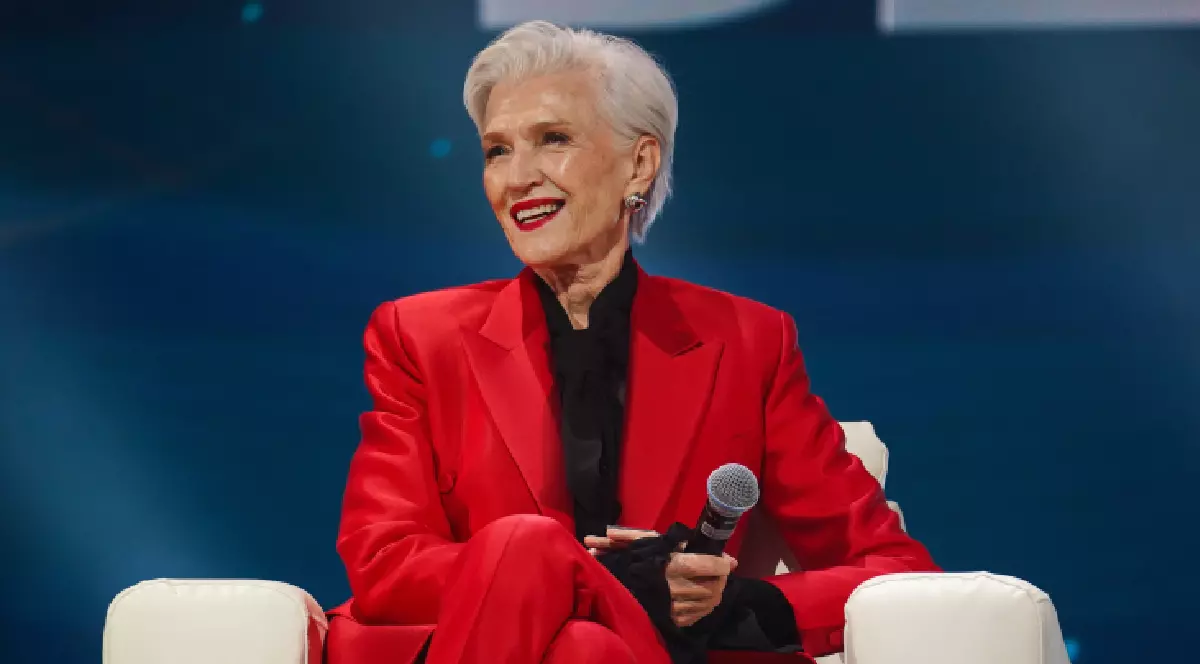 Maye Musk, mama lui Elon Musk, îmbrăcată într-un costum roșu, având un microfon în mână