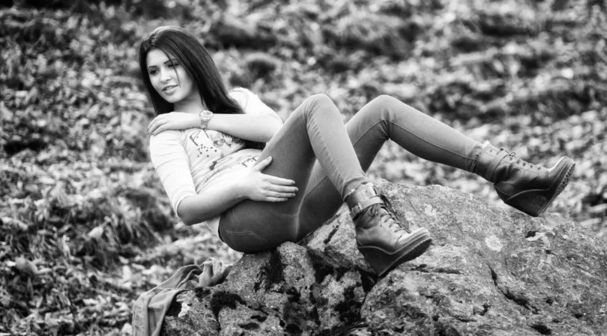 În imagine este o femeie tânără care stă relaxată pe o stâncă, într-un decor natural de toamnă. Poartă bocanci, pantaloni strâmți și un tricou cu mânecă trei sferturi, iar expresia ei este calmă și contemplativă.