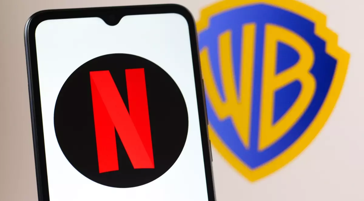 În imagine se vede un telefon pe al cărui ecran apare sigla Netflix, cu litera roșie „N” pe fundal negru. În spatele telefonului, neclar, se distinge logo-ul Warner Bros., sugerând contextul unui parteneriat sau al unei achiziții între cele două companii.