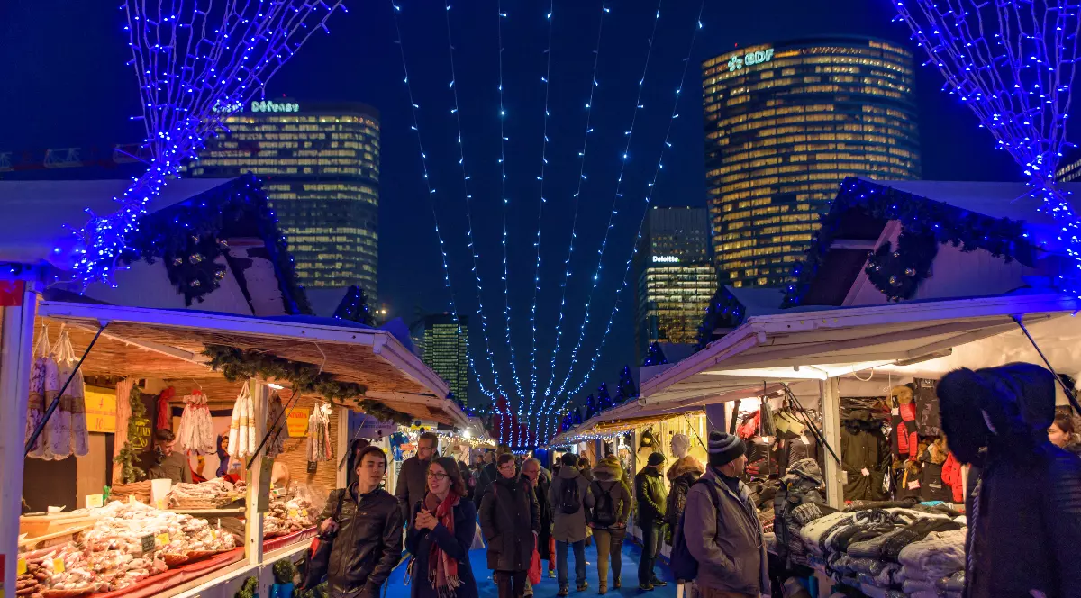 Imaginea surprinde o piață de Crăciun aglomerată, luminată intens de instalații albastre suspendate deasupra aleii principale. De o parte și de alta se aliniază căsuțe de lemn decorate festiv, unde comercianții vând produse tradiționale, mâncare și articole de iarnă, în timp ce vizitatorii se plimbă printre standuri într-o atmosferă animată de sărbătoare.