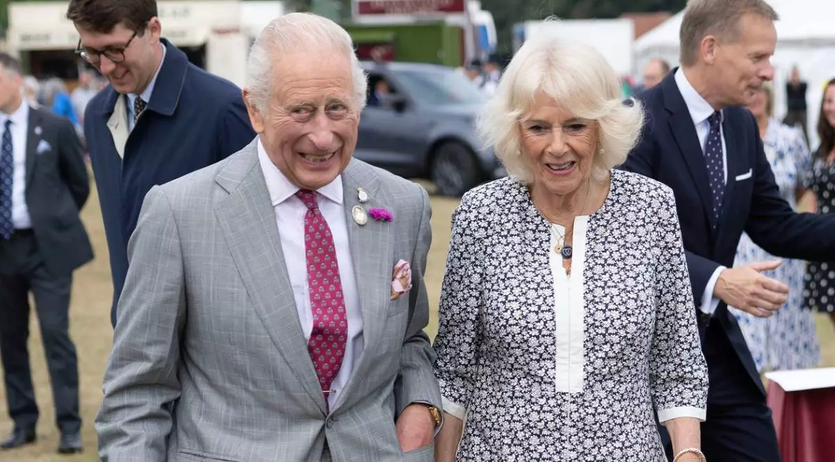 Regela Charles, stanga, in costum gri si cravata rosie, alaturi de regina Camila, dreapta, in rochie cu imprimeu marunt si guler alb, la un eveniment in aer liber