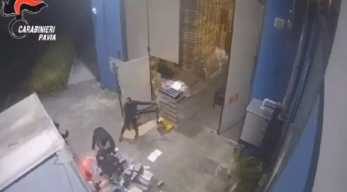 O imagine surprinsă de o cameră de supraveghere, noaptea, arată o persoană îmbrăcată în haine întunecate care pătrunde într-un depozit, dezvăluind cutii stivuite în interior.