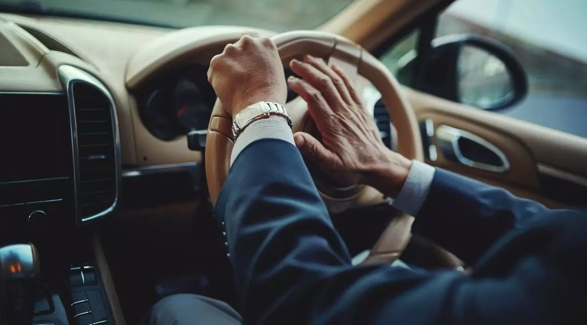 Mâinile unui bărbat îmbrăcat la costum și care poartă ceas se văd pe volanul unei mașini de lux