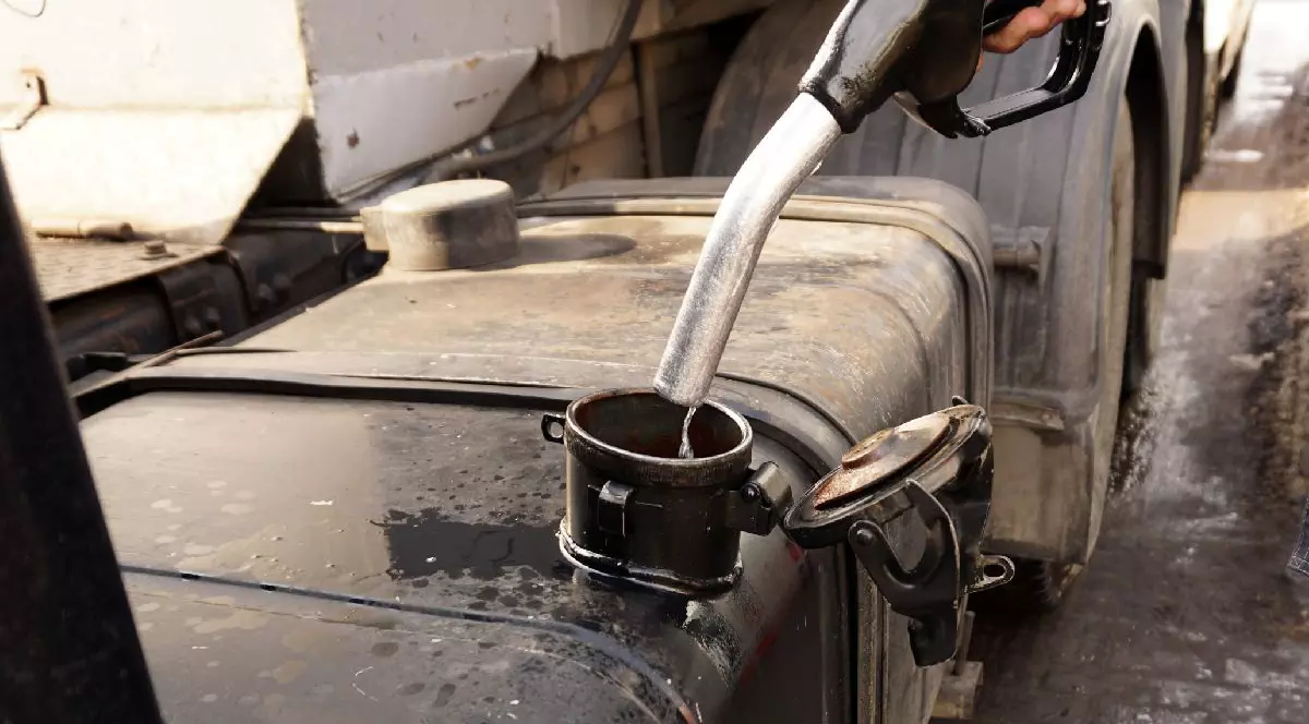 Prim-plan al unei persoane care toarnă motorină dintr-o pompă de combustibil neagră într-un rezervorul unui camion.În fundal se văd șasiul metalic și anvelopele vehiculului greu.