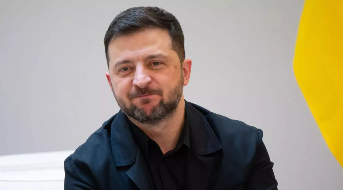 În imagine Volodimir Zelenski, presedintele Ucrainei, îmbrăcat într-un costum închis la culoare, cu barbă scurtă și păr tuns scurt, așezat într-un decor sobru. În fundal se distinge parțial un steag galben, iar expresia sa este calmă și concentrată, sugerând un cadru oficial.