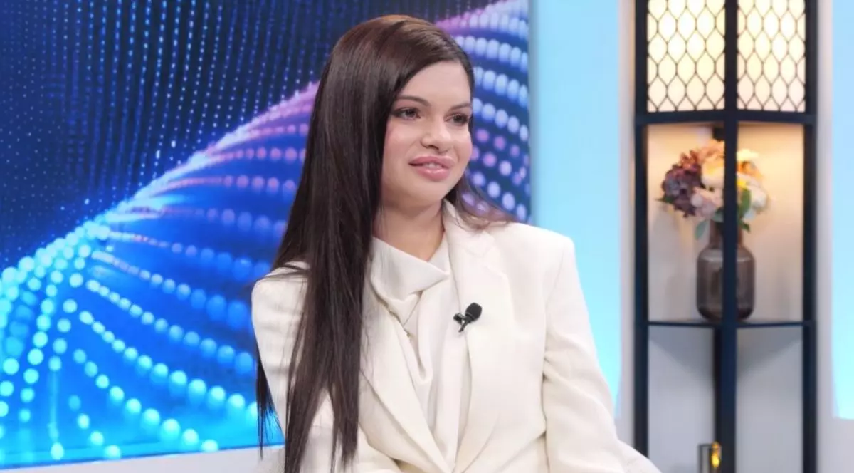 Ana-Maria Borlovan, îmbrăcată cu un costum alb, cu sacou alb, cu părul șaten lung, în timp ce stă pe scaun, iar pe fundal se vede un ecran albastru și o floare pe un raft