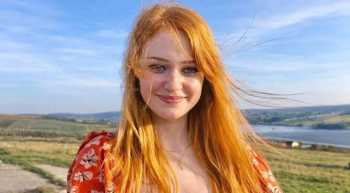 Andreea Anita, cu parul portocaliu, desfacut, zburat de vant, intr-o rochie rosie cu flori albe, zambeste intr-o zi insorita, se vede un cer albastru, in spate dealuri verzi și o apa, Andreea, din Falticeni, are nevoie de 2.3 milioane de dolari pt un tratament in SUA