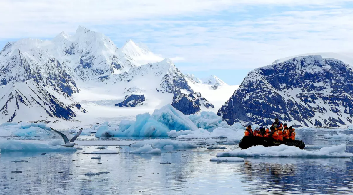 Un grup de turiști într-o barcă gonflabilă în Arctica, stau în barcă în timp ce ghidul le arată muntele de gheață din spatele lor, acesta este complet acoperit de zăpadă