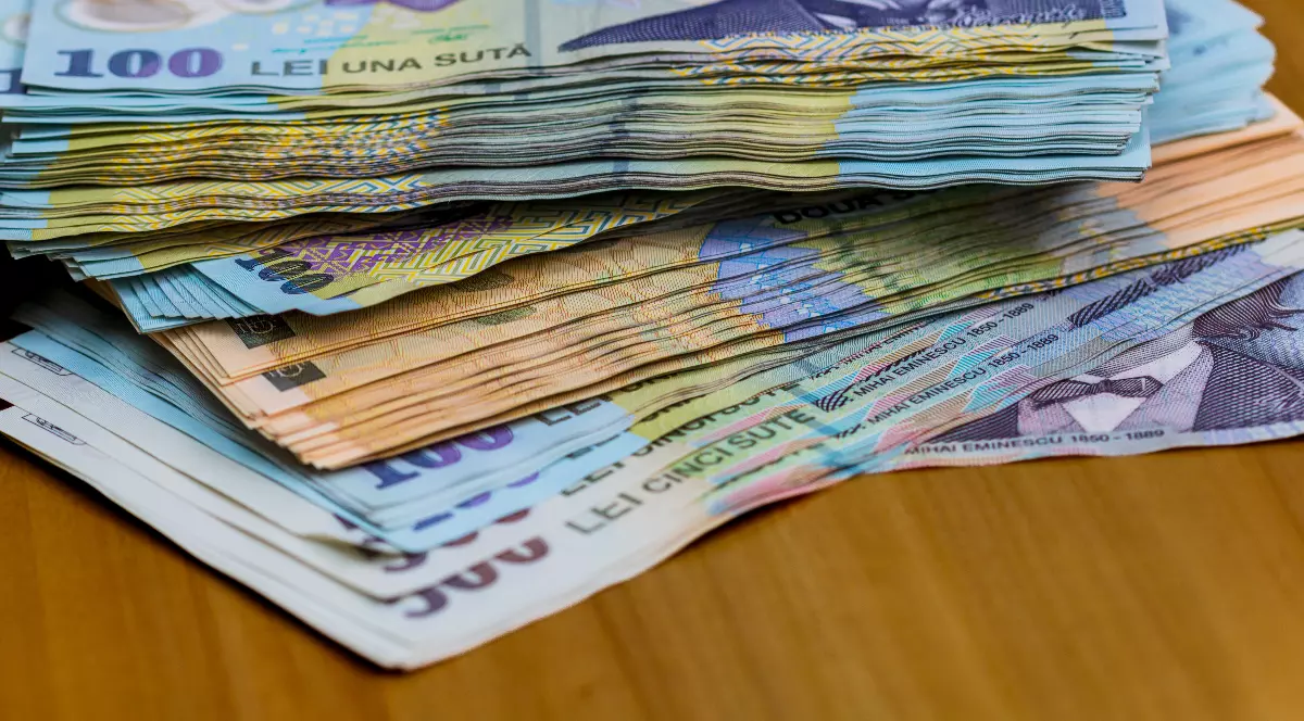 Imaginea prezintă mai multe teancuri de bancnote românești, de diferite valori, așezate pe o suprafață de lemn. Culorile distincte și portretele de pe bancnote sugerează sume importante de bani și un context financiar sau economic.