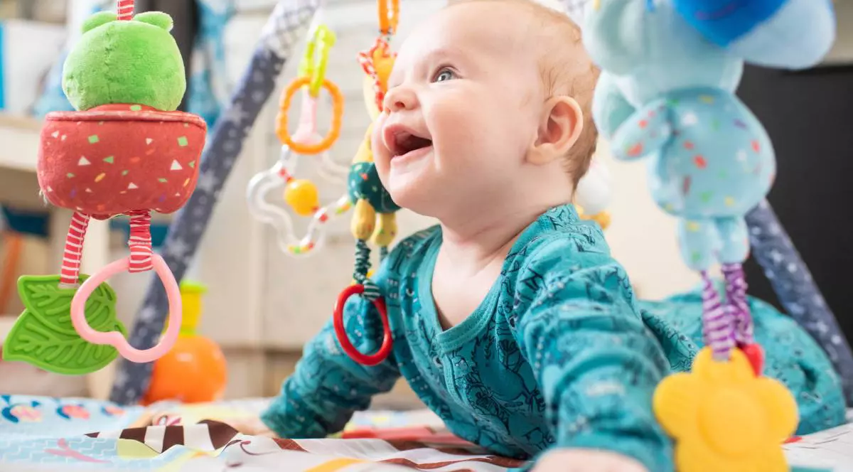 Bebelus intr-o blluza verde, zambeste in timp ce sta pe burta pe o salteluta de activitati, se vad multe jucarii specifice varstei, colorate, atarnate,