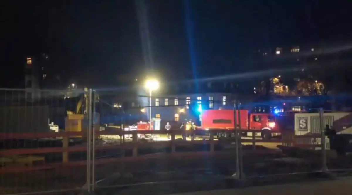 Imagine nocturnă surprinsă pe un șantier. În spatele unui gard metalic se văd utilaje oprite și o autospecială roșie de intervenție, cu girofarurile albastre aprinse. Câteva persoane în veste reflectorizante stau lângă vehicul. În fundal apar clădiri de locuințe cu ferestre luminate. Fotografia surprinde o intervenție a Interventia autorităților din Aachen, după descoperirea unei bombe de 250 de kilograme pe un șantier, și evacuarea a peste 6.000 de persoane, 21 ianuarie 2026