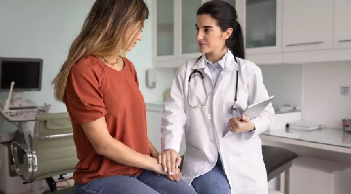 O femeie cu parul lung, imbracata intr-un tricou maro, stă de vorbă cu o femeie medic, imbracata in halat si cu stetoscop la gat. Medicul o tine de mana. Cancer col uterin. Vaccin HPV. Foto: Shuterrstock