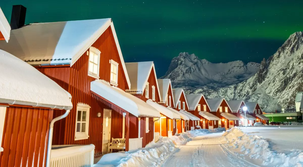 Case îmbrăcate în lemn de culoare roșie, în partea stângă, iar pe centru un drum acoperit de zăpadă, iar pe fundal cerul pe timp de noapte cu aurora boreală