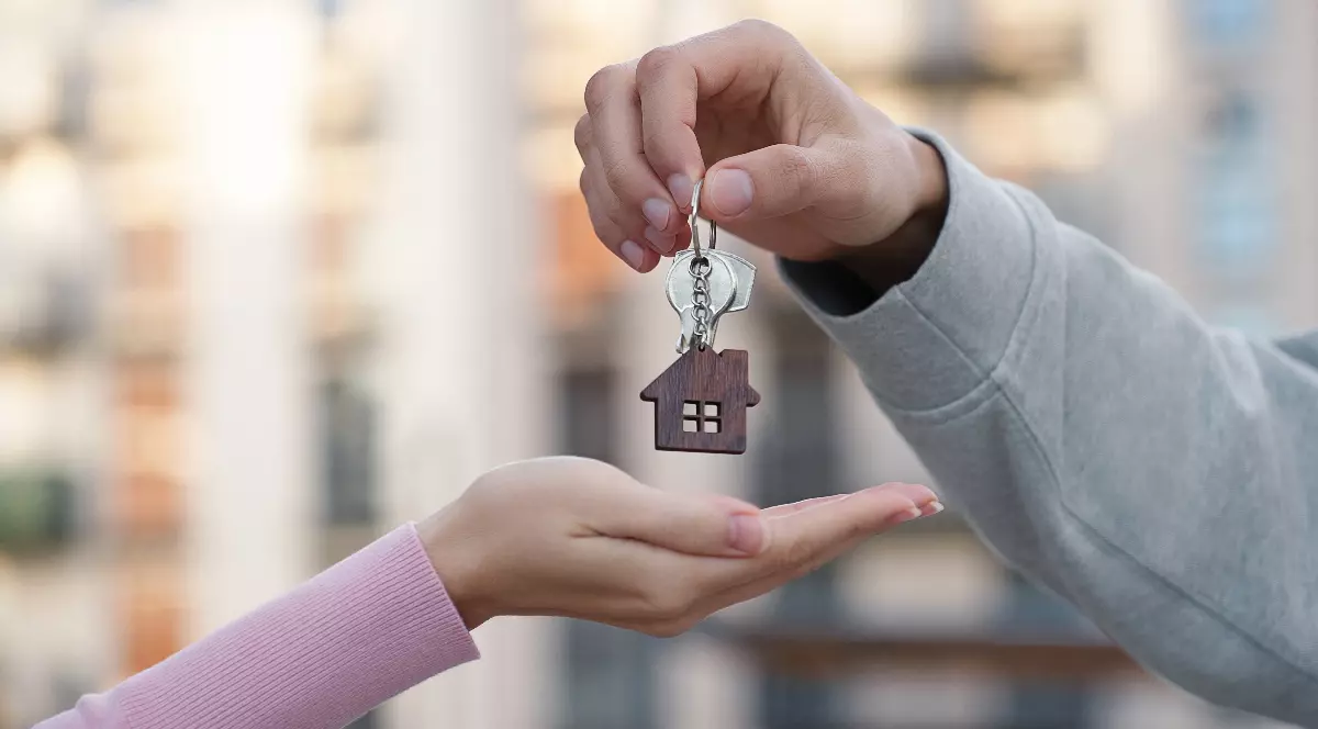 O mână a unui bărbat oferă cheile, care conțin și un breloc cu o casă, unei femei