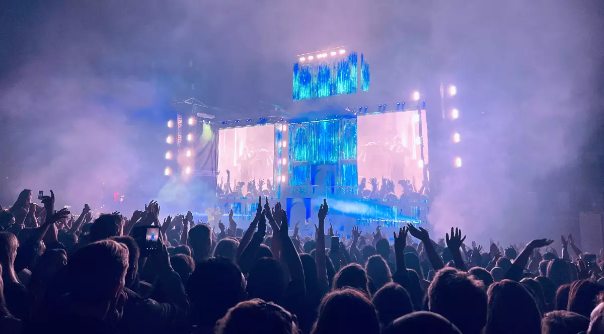 Imaginea surprinde un concert în aer liber, cu o scenă mare iluminată și ecrane uriașe, în fața unei mulțimi numeroase care ridică mâinile și filmează momentul. Atmosfera este una energică, accentuată de lumini, fum de scenă și entuziasmul publicului.