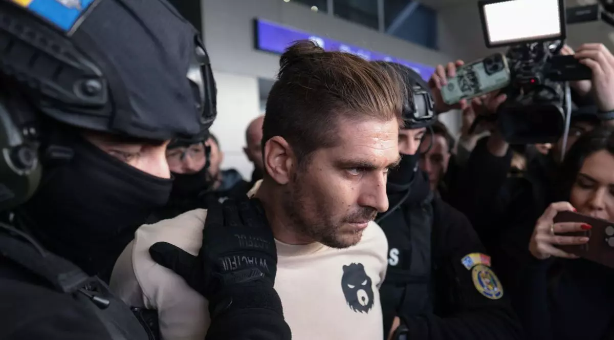 Cosmin Zuleam este fotografiat din semiprofil, avand o tunsoare în care partea de calotă are părul mai lung, iar lateralele sunt tunse scurt. Mâna unui polițist este pe pieptul lui iar din directia opusă se văd pe fundal jurnaliștii care îl fotografiază.