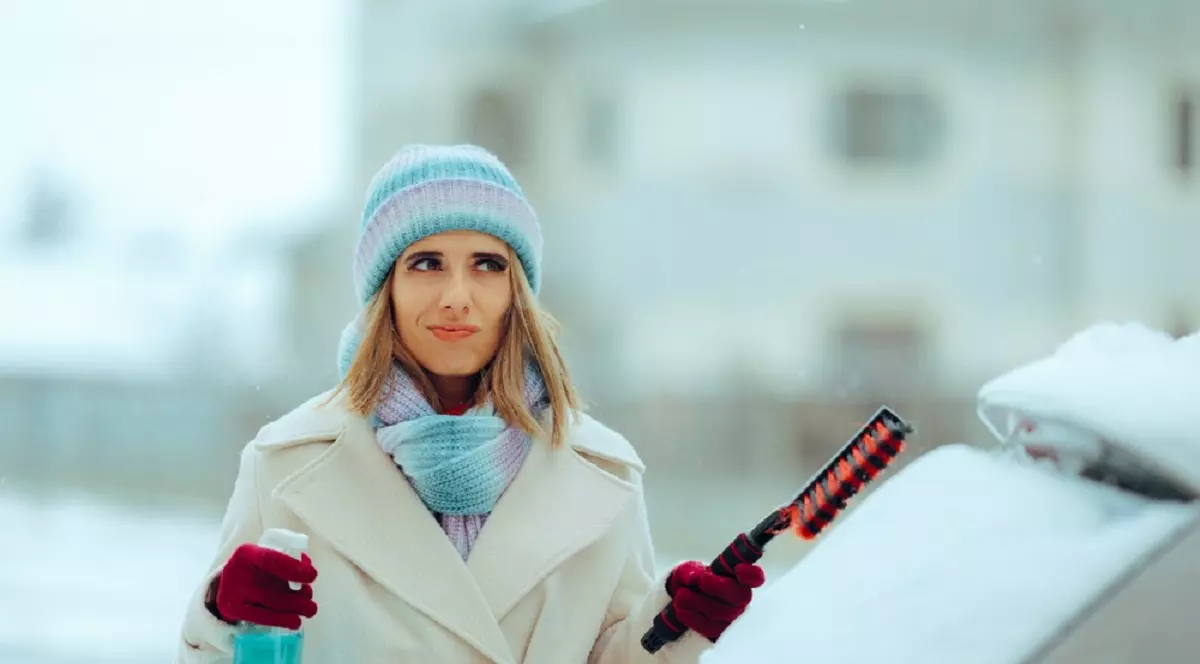 Cum dezgheți ușor portiera și parbrizul când este ger - O femeie echipata de iarna, cu o perie si un lichid de parbriz se pregateste sa curete masina de zapada