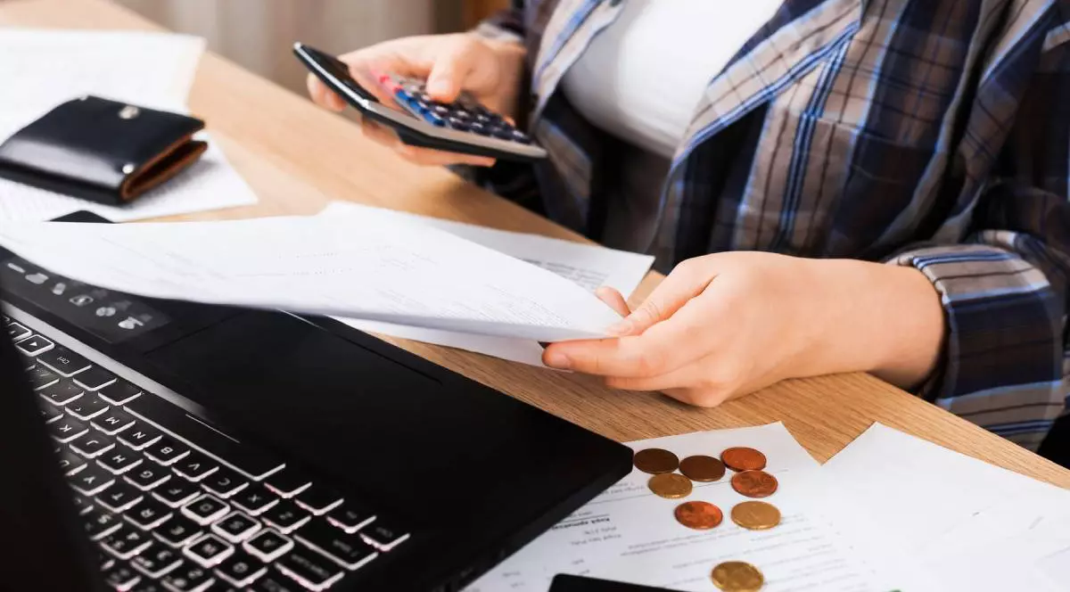 Femeie verificând facturi și folosind un calculator lângă un laptop. Concept de buget gospodăresc cu monede, card de credit și documente pe masă.