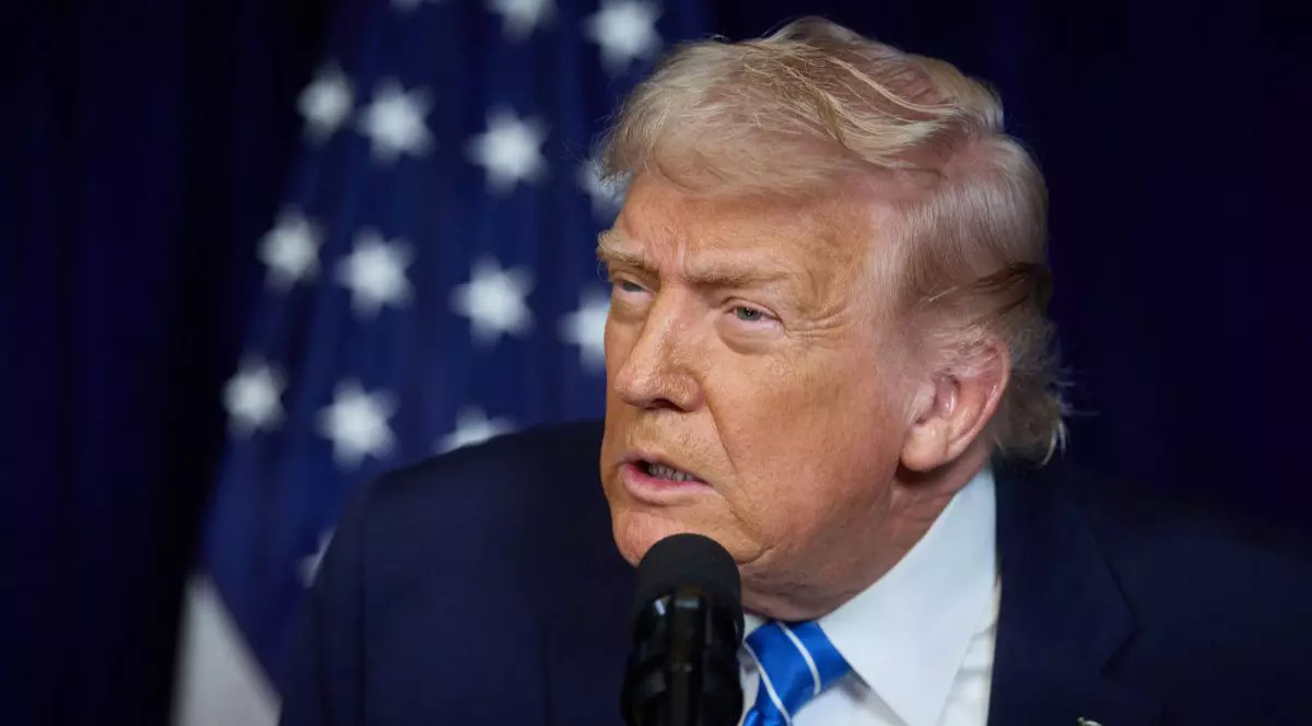 Portret Donald Trump, in sacou negru, camasa alba, cravata cu dunci albastre si insigna in piept, surprins in timp ce vorbeste la microfon, in spate se vede steagul SUA