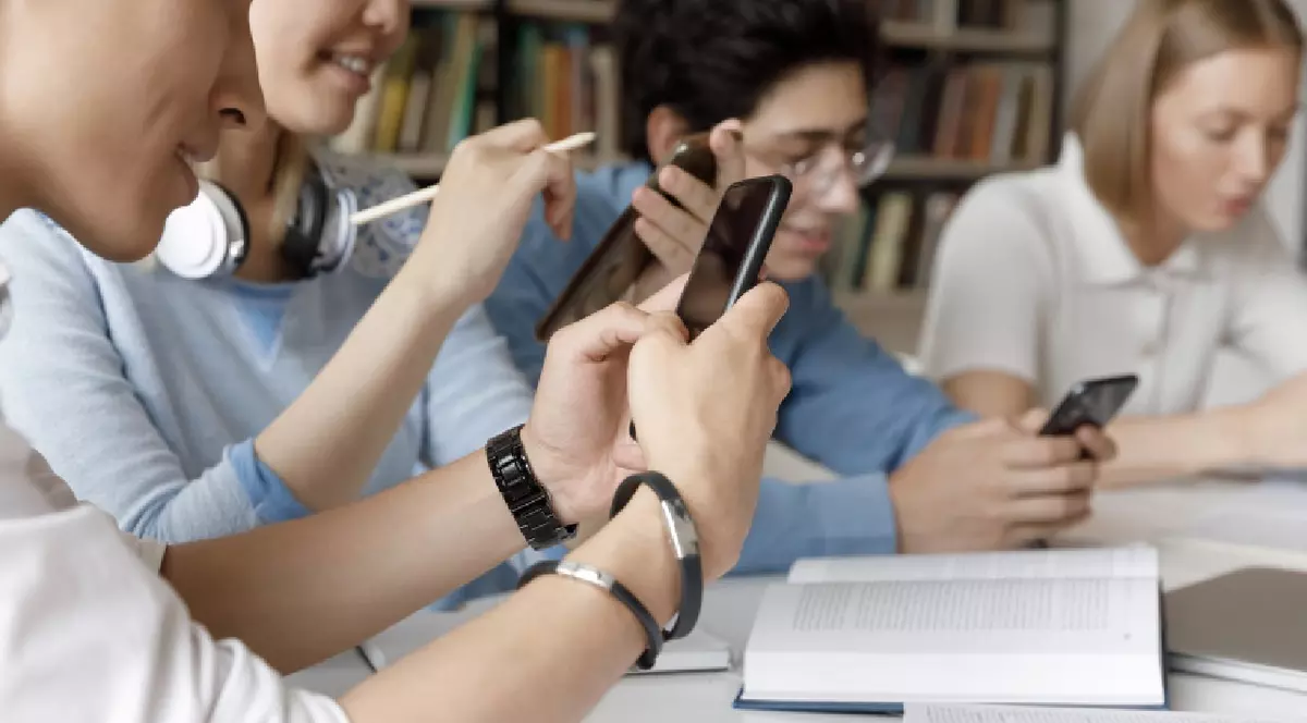 Grup de tineri studenți din generația Z care navighează pe internet în biblioteca campusului, folosind smartphone-uri la masa de lucru cu cărți,