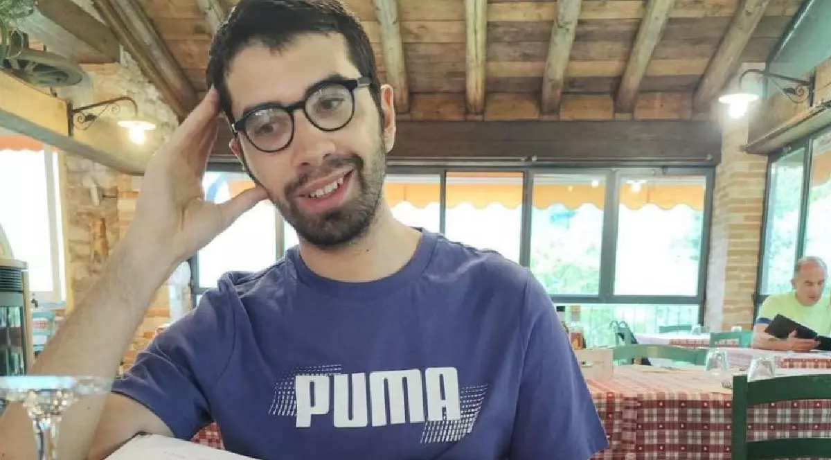 Fotografie cu un tânăr cu ochelari și barbă, îmbrăcat cu un tricou albastru Puma, zâmbind într-un restaurant rustic.