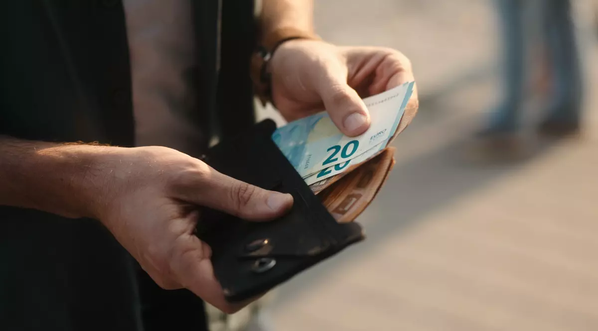 Imaginea surprinde o persoană care scoate o bancnotă de 20 de euro dintr-un portofel negru, ținut în mână.