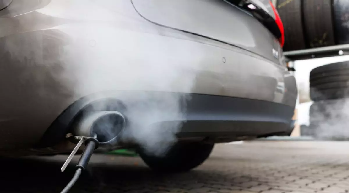 Fotografie cu țeava de eșapament a unei mașini argintii care emite fum alb dens, cu anvelope stivuite în fundal.