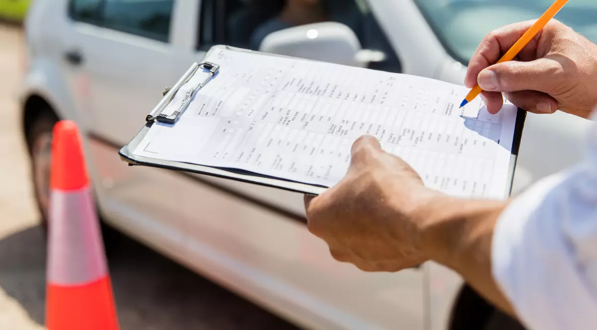 Un examinator auto care scrie pe o foaie. În fundal se observă o mașină albă.