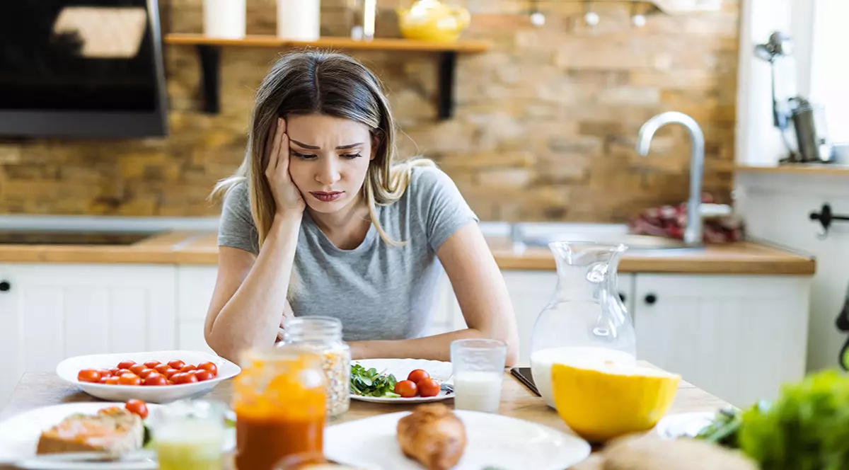O femeie îmbrăcată într-un tricou gri, suferind de depresie, stă în fața unei mese pline.