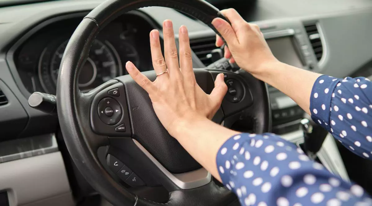 Imagine neutra cu mainile unei femei cu o bluza albastra cu buline albe pe volanul masinii, cu mana dreapta apasa claxonul