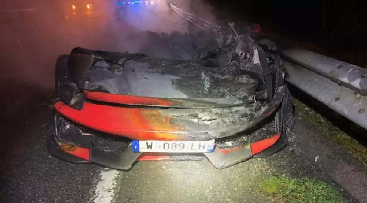 Fotografie cu un Ferrari roșu cuprins de flăcări pe un drum, noaptea, lângă un parapete, cu lumini de urgență în fundal.