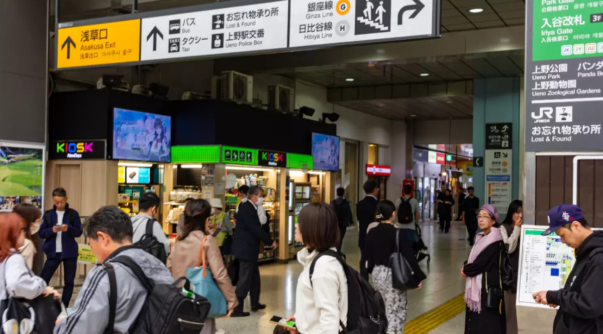 Mai mulți turiști într-o gară din Tokyo, în timp ce urmăresc panourile și afișajul trenurilor și își cară bagajele