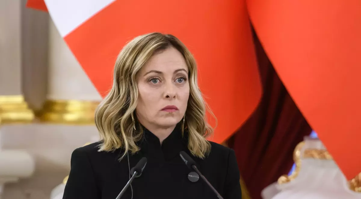 Imaginea o surprinde pe o femeie aflată la un pupitru oficial, cu microfoane în față, având o expresie serioasă și concentrată. Fundalul cu elemente decorative și culori puternice sugerează un cadru instituțional sau o conferință oficială.