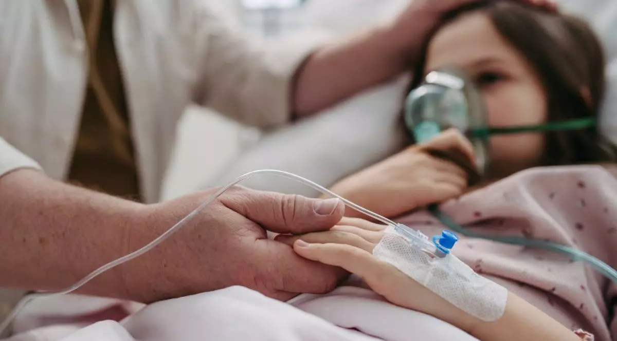 Mana unui barbat in halat alb tine de mana o fetita, aflata pe patul de spital, cu o masca de oxigen pe fata,