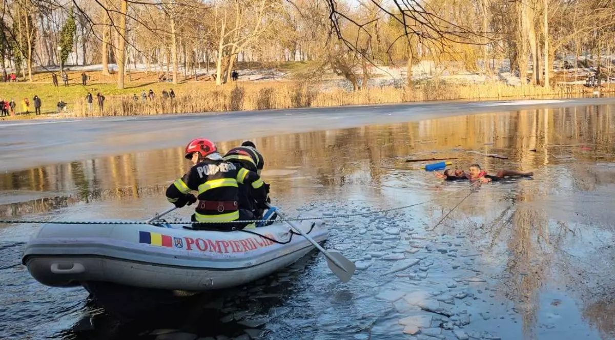 Interventie fetita cazuta in apa Parc Romanescu Craiova, 17 ianuarie 2026,in imagine se vede o barca pneumatica cu doi pompieri, vaslesc pe apa inghetata, la cativa metri de ei doi barbati si o fetita in apa, se vad copaci cu zapada pe langa apa, e o zi insorita de iarna