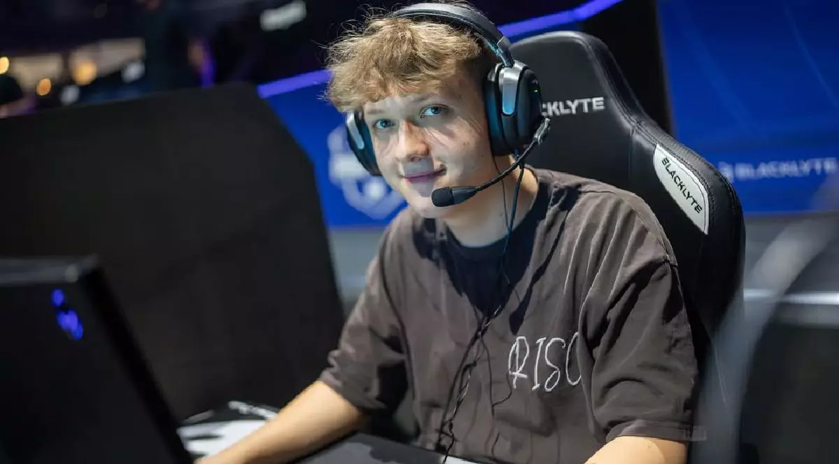 Fotografie cu un tânăr jucător cu părul creț, purtând căști, tricou negru și microfon, așezat pe un scaun de jocuri, în fața unui computer.