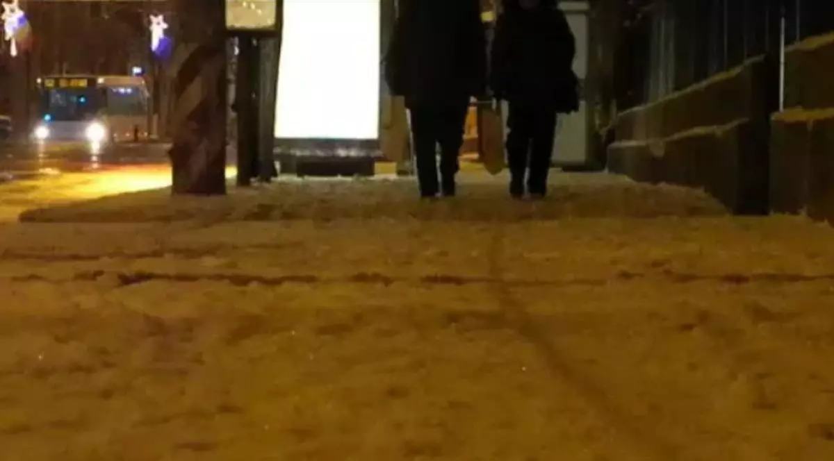O stradă acoperită de zăpadă pe care merg doi oameni fotografiați cu spatele, sunt în dreptul unei stații de autobuz, este seară, iar pe partea cealaltă se vede un autobuz în trafic cu luminile aprinse, Cluj Napoca, ianuarie 2026