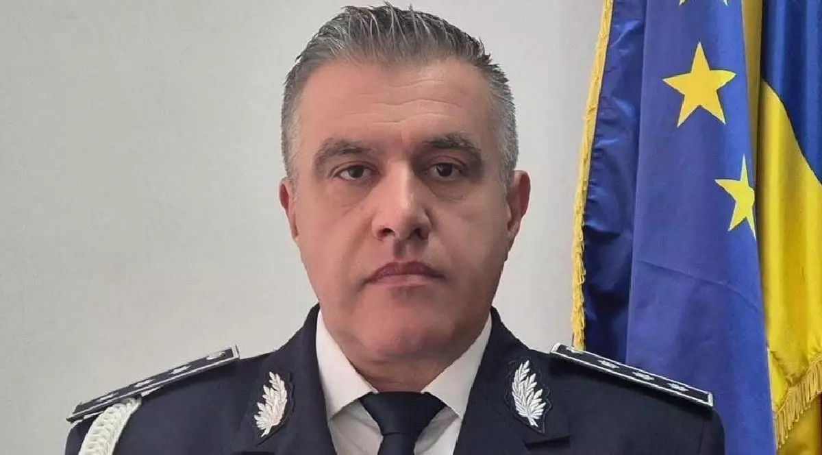 Fotografie a unui bărbat de vârstă mijlocie, cu părul scurt și cărunt, îmbrăcat în uniformă închisă la culoare, cu epoleți argintii și cravată neagră, stând în fața unui steag.