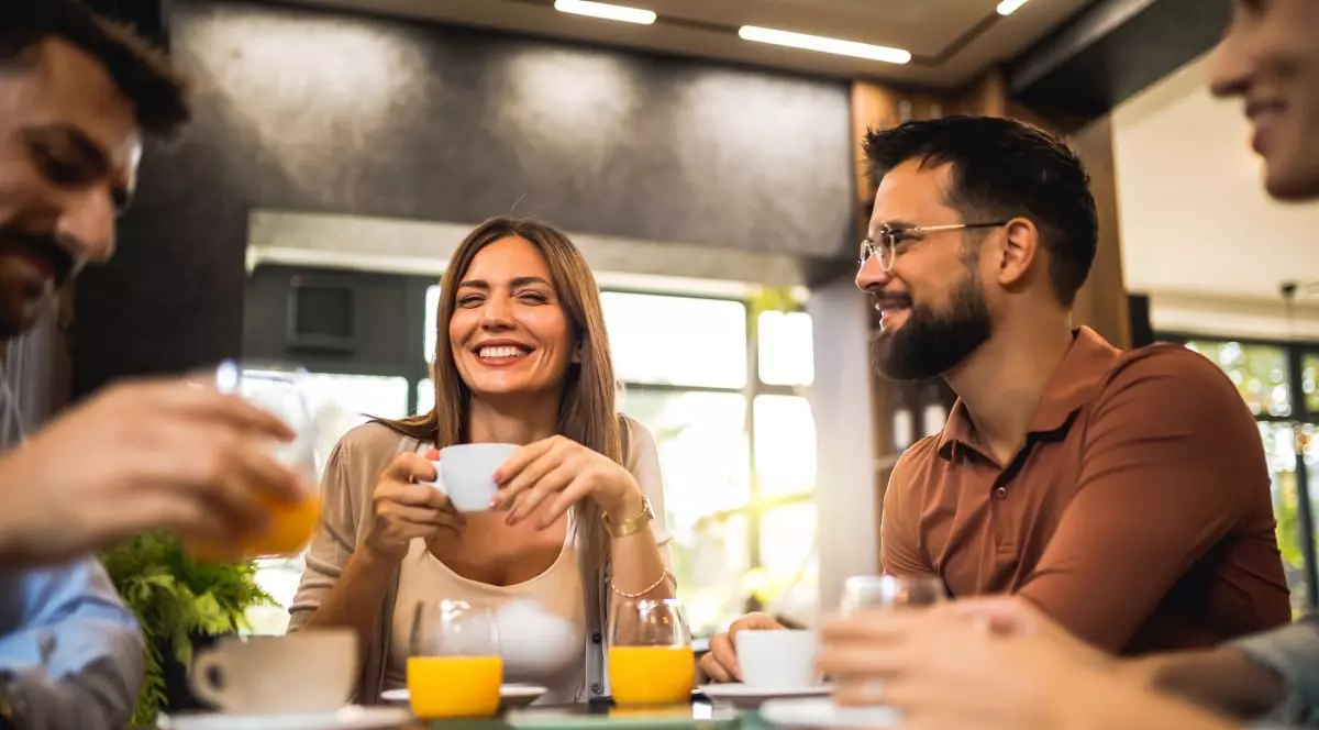 Un grup de prieteni este la o cafenea. Un centru este o femeie cu o bluza albă care are o cană albă în mână și râde, iar în jurul ei sunt trei bărbați