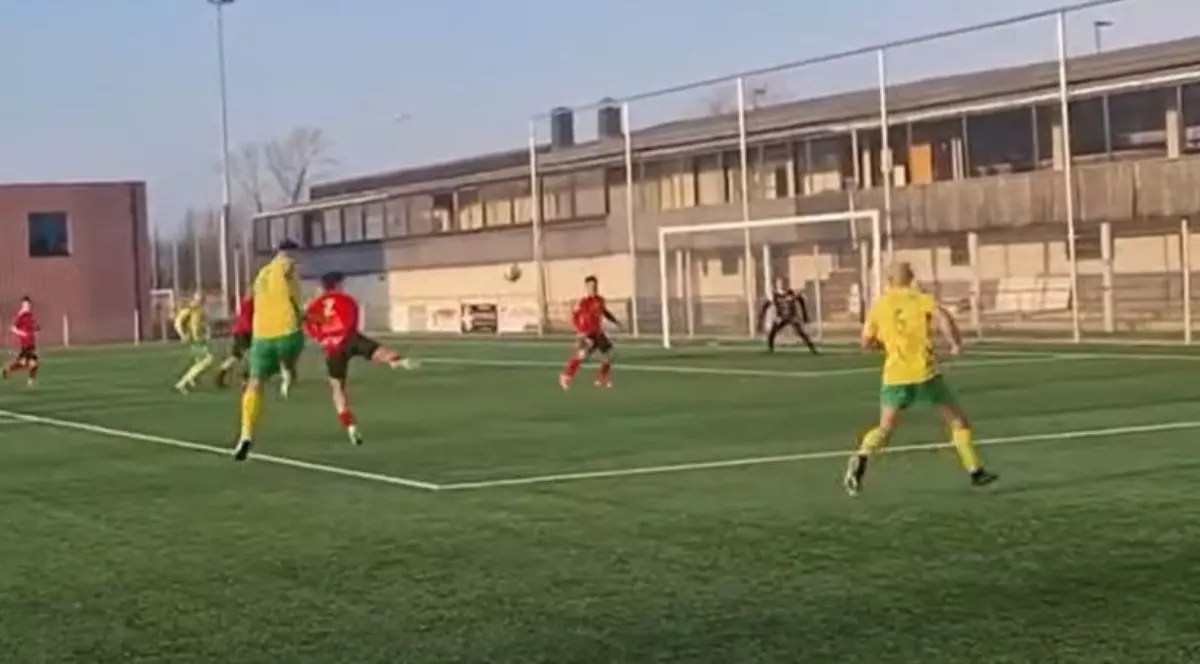 o fază dintr-un meci de fotbal desfășurat pe un teren cu gazon artificial verde, delimitat de linii albe clare. În centrul acțiunii, mai mulți jucători sunt dispersați pe teren: unii poartă echipamente galbene cu șorturi verzi, în timp ce adversarii lor sunt îmbrăcați în tricouri și șorturi roșii. Mingea este vizibilă în aer, în apropierea careului de pedeapsă, unde un jucător în roșu pare să fi executat o lovitură. În fundal, se află poarta de fotbal cu plasă albă, în fața căreia este poziționat portarul, iar mai departe se întinde o clădire lungă cu un etaj, prevăzută cu numeroase ferestre mari de sticlă și un acoperiș plat