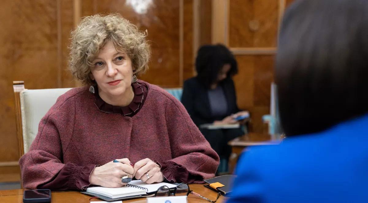 Vicepremierul Oana Gheorghiu, în timpul unei ședințe la Palatul Victoria, poartă un palton vișiniu și stă așezată pe scaun, la masă