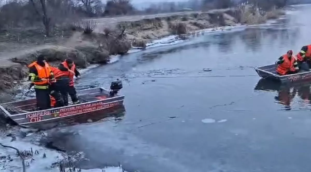 In imagine se vad doua barci ale Isu, una la mal si una pe apa, mai multi salvatori care cauta persoane cazute in raul Olt, iarna, malul este inghetat, ziua este intunecata si rece