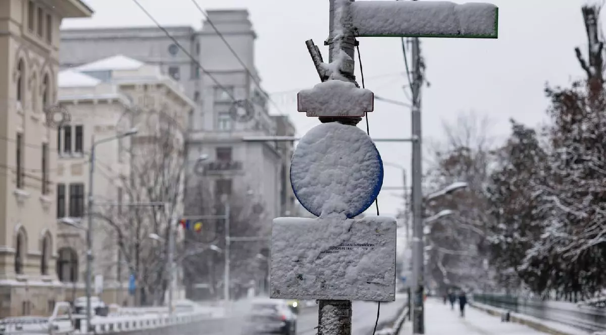 Fotografie cu o stradă acoperită de zăpadă, cu un stâlp de iluminat și semafor, clădiri în fundal și câteva persoane mergând pe trotuar.