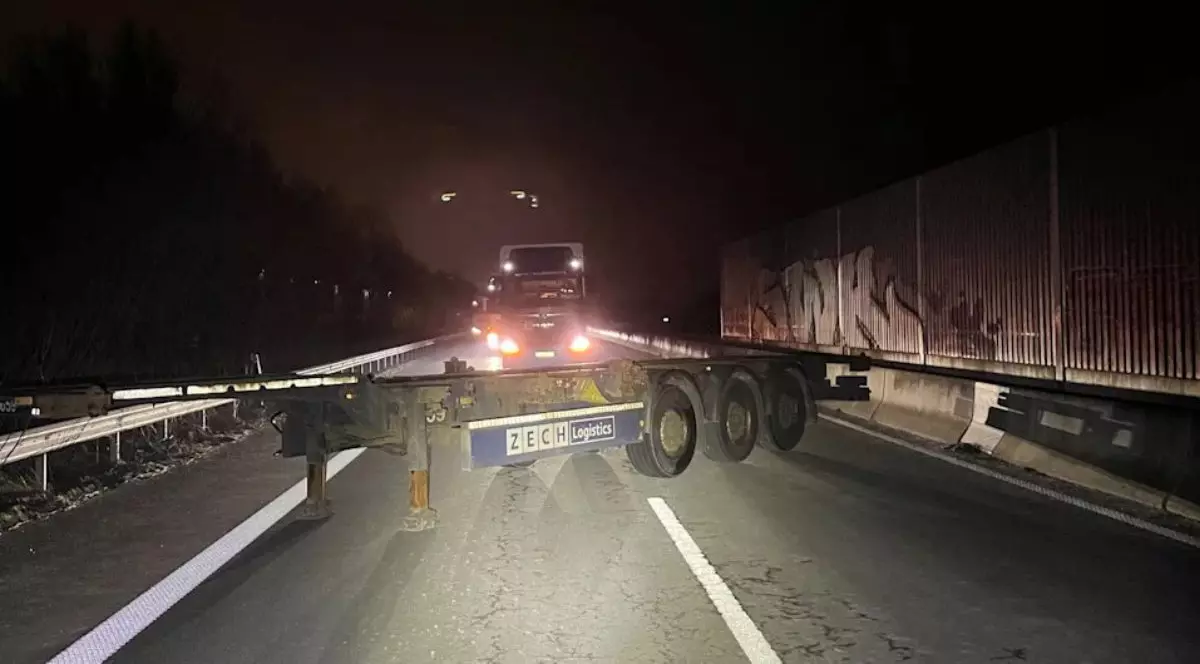 o secțiune de autostradă unde o semiremorcă lungă, de culoare închisă, este poziționată perpendicular pe direcția de mers, blocând ambele benzi de circulație. Semiremorca are vizibil logoul „ZECH Logistics” pe o porțiune albastră și este sprijinită în partea stângă pe picioarele metalice de susținere, în timp ce în partea dreaptă se observă cele trei axe cu roți. În plan secund, un cap tractor cu farurile aprinse este oprit în fața obstacolului, lumina acestuia reflectându-se pe carosabil și pe elementele metalice ale remorcii