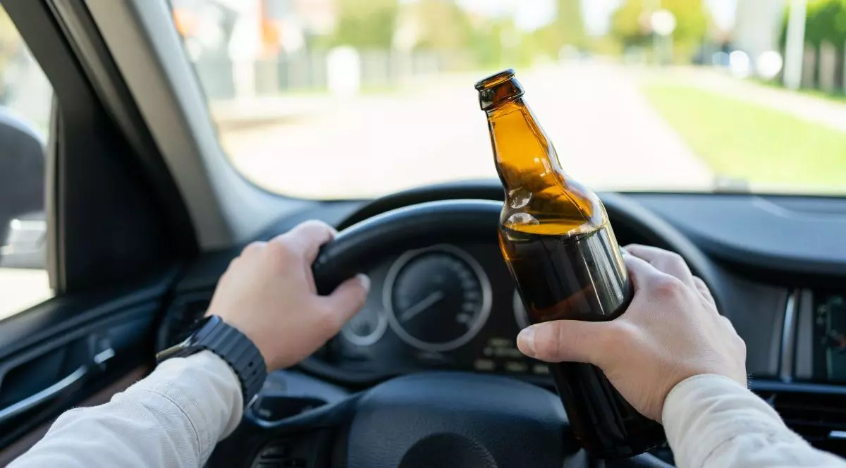 Un bărbat conduce o mașină, ține o sticlă de alcool în mână și este cu mâinile pe volanul mașinii