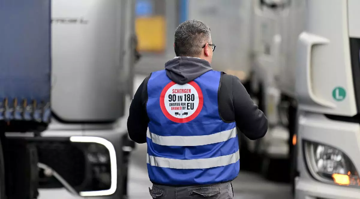 Un bărbat de vârstă mijlocie, cu părul scurt și cărunt, îmbrăcat cu o vestă albastră stă între două camioane mari.