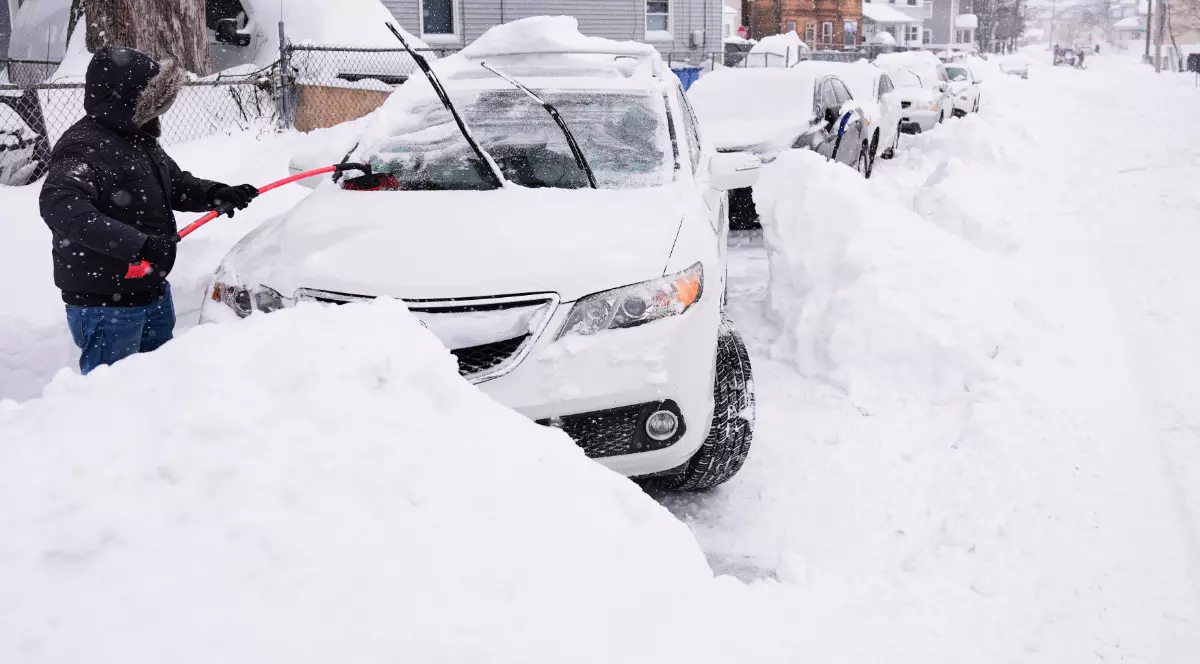 O persoană îmbrăcată gros curăță zăpada de pe un autoturism acoperit aproape complet, într-o zonă rezidențială. Strada este blocată de nămeți înalți, iar mașinile parcate sunt îngropate în zăpadă, sugerând impactul sever al furtunii.