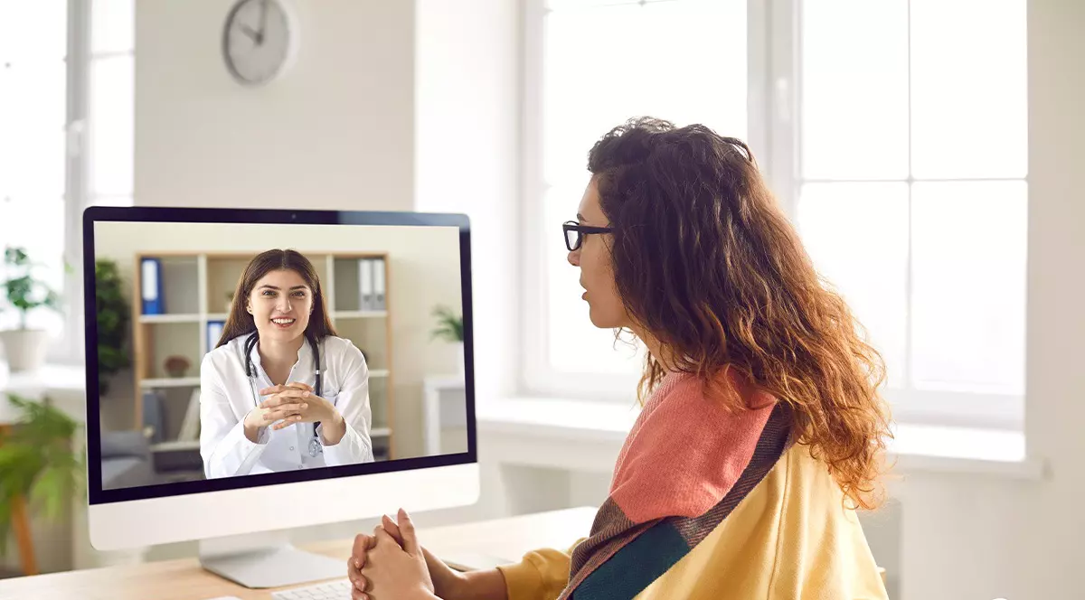 O femeie cu parul lung si ochelari cu un pulover galben sta in fata computerului di priveste la o femeie in halat alb, medic.Telemedicina nu e decontata la medicul de familie. Teleconsultatie. Romani din rural fara acces la servicii medicale. Foto; Shutterstock