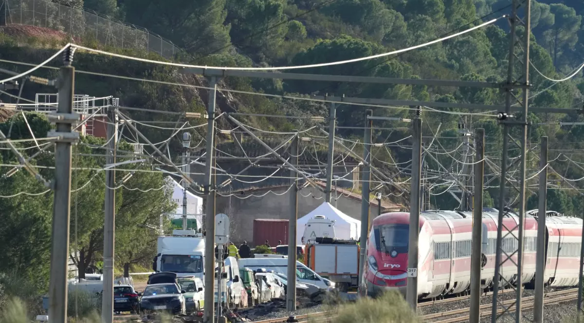 Imaginea arată un tren de mare viteză oprit pe o linie ferată, într-o zonă deluroasă împădurită, unde sunt prezente vehicule și corturi de intervenție. Contextul sugerează un accident feroviar, cu traficul blocat și echipe de urgență aflate la fața locului.