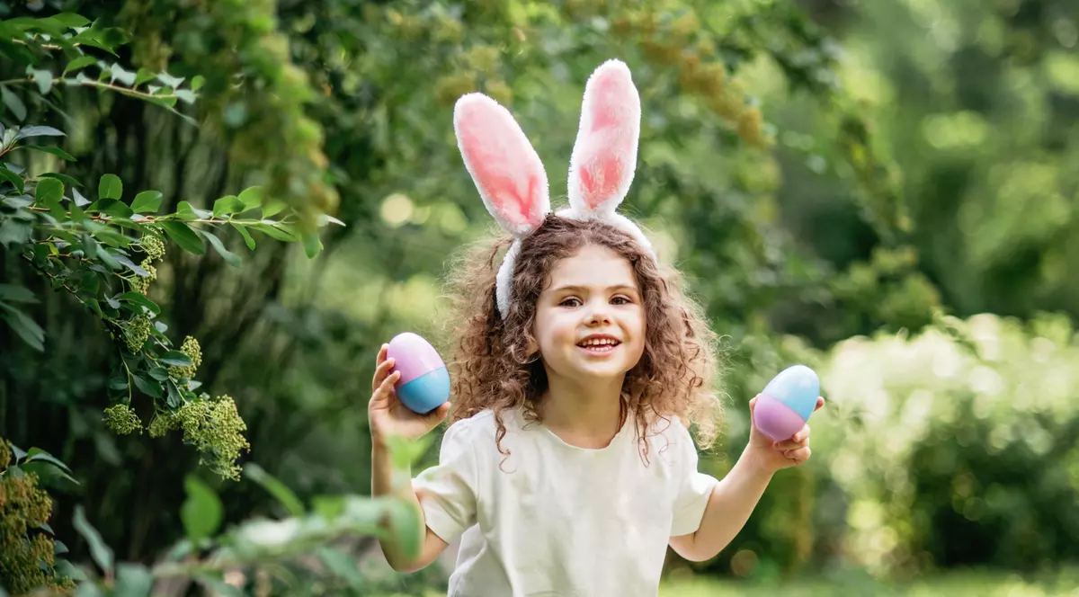 O fetiță are urechi de iepuraș și ține în mâini câte un ou colorat de Paște